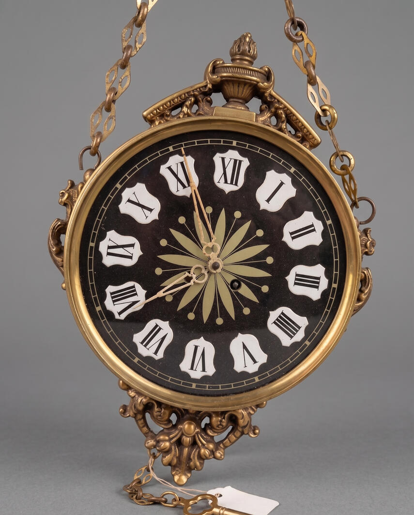 Vintage ornate German brass hanging wall clock in Classical Revival style with a black face and engraved gold center.
